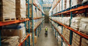 Large and high racking of pallet storage in Beeston 3PL warehouse with worker moving goods on pump truck. 