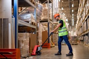 Warehouse worker moving pallet storage with a pump truck down warehouse aisle. 